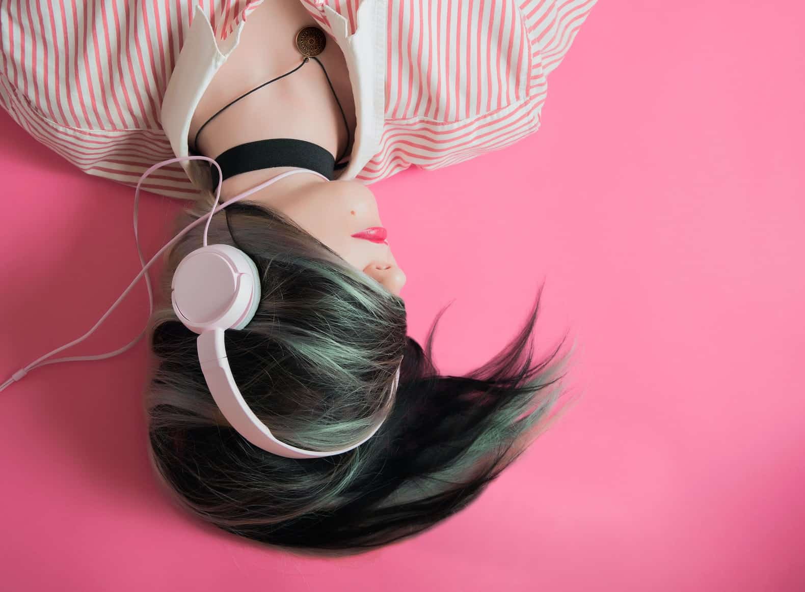 pale skinned woman with green and black hair obscuring her face, lying on a pink floor with headphones on listening to podcasts