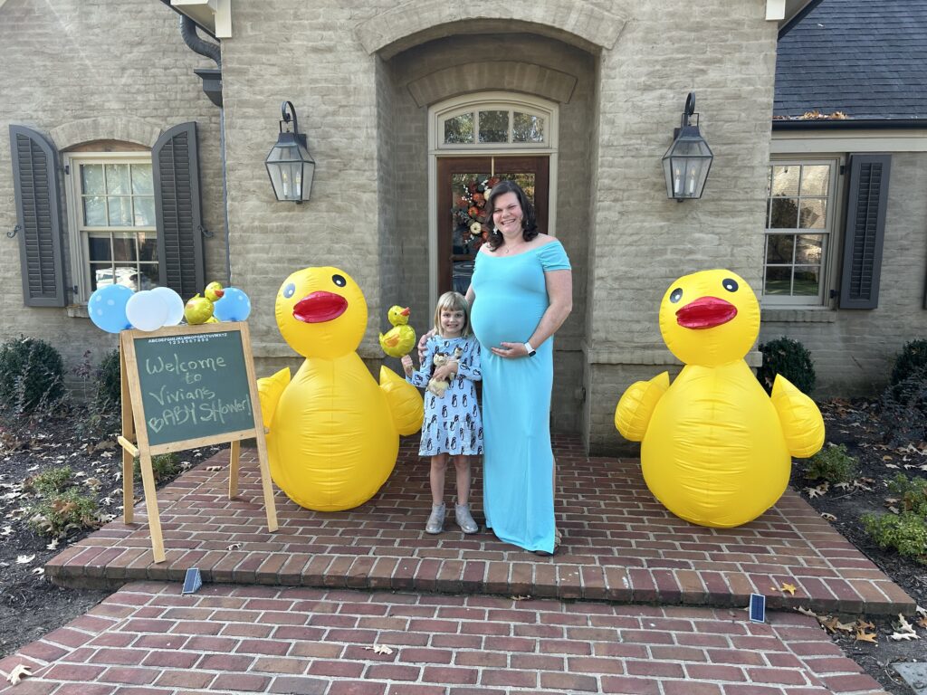 Little girl and pregnant woman standing in front of a front door with two blow up ducks next to them and a sign that says Welcome to Vivian's baby shower