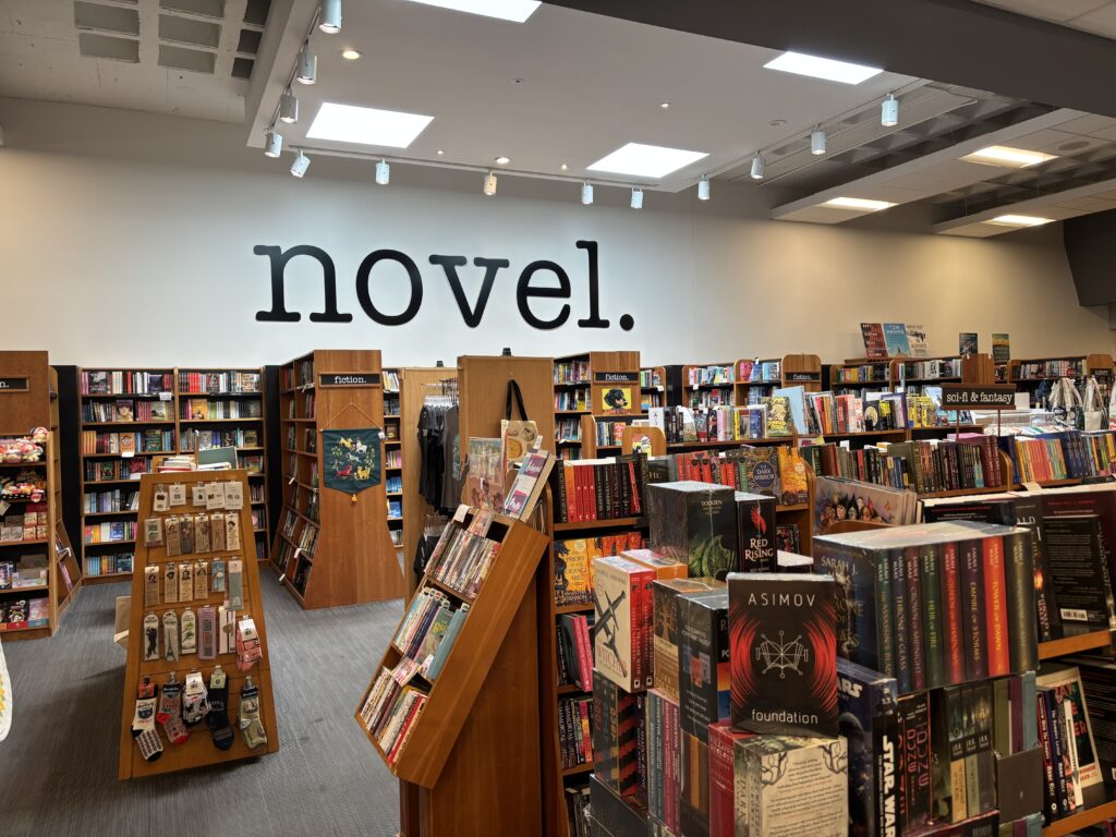 Many brown bookshelves with may different kinds of books on it, with the big black letter sign "Novel" above it on the wall